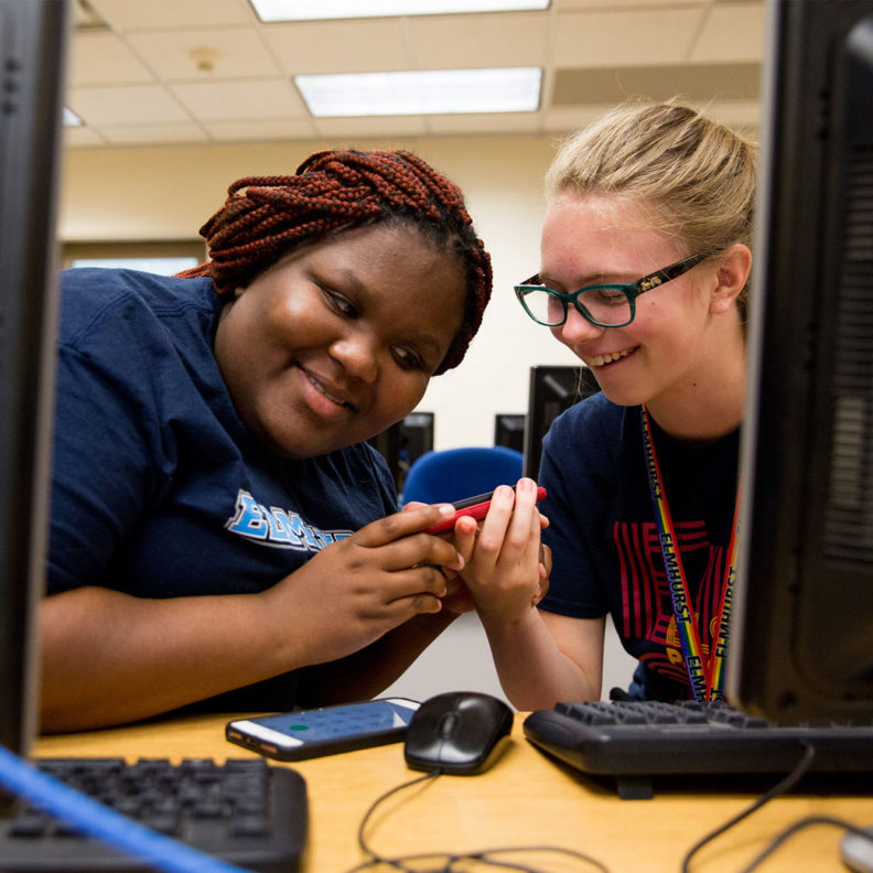 埃尔姆赫斯特大学的一名黑人女学生和一名白人女学生在电脑终端之间相视一笑。beplay体育飞翔下载